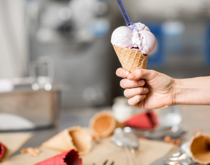 Holding waffle cone with ice cream indoors Holding waffle cone with ice cream at the kitchen with food ingredients and ice cream maker machine on the background
